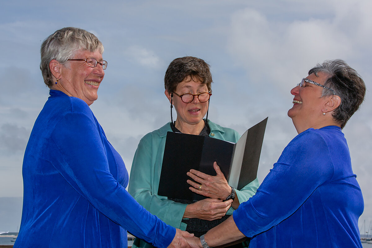 Marj McCann and Carole Smith, dressed all in blue, hold hands while an officiant marries them on the beach in Provincetown, Massachusetts.